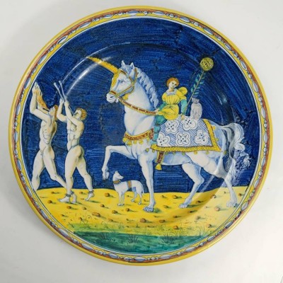 TOSKANISCHER WANDTELLER, Majolika, Kopie des 20. Jh. nach einem von Jacopo di Stefano Schiavone (um 1490 - nach 1576) bemalten Teller, entstanden 1514, der sich heute im Fitzwilliam Museum in Cambridge befindet. Die Darstellung zeigt einen Ausschnitt aus einem Triumph Caesars, nämlich Caesars Pferd. Auf der linken Seite sind zwei nackte Musiker mit Glocken um die Knöchel zu sehen, die Doppelpfeifen in die Höhe halten und blasen. Hinter ihnen steht ein Hund mit Glöckchen am Halsband und einem 