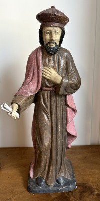 Anonym (19. Jahrhundert) Heiligenfigur, um 1850, Holz. Es handelt sich um eine Darstellung eines Heiligen, eines Mönchs oder eines biblischen Gelehrten. Die Figur trägt eine lange Robe in dunklem Braun mit einem rötlichen Umhang, was typisch für Mönche oder Gelehrte aus dem Mittelalter oder der Renaissance ist. In der einen Hand hält die Figur eine Schriftrolle, in der anderen ist die Hand auf die Brust gelegt - ein Zeichen von Hingabe, Gebet oder Weisheit.